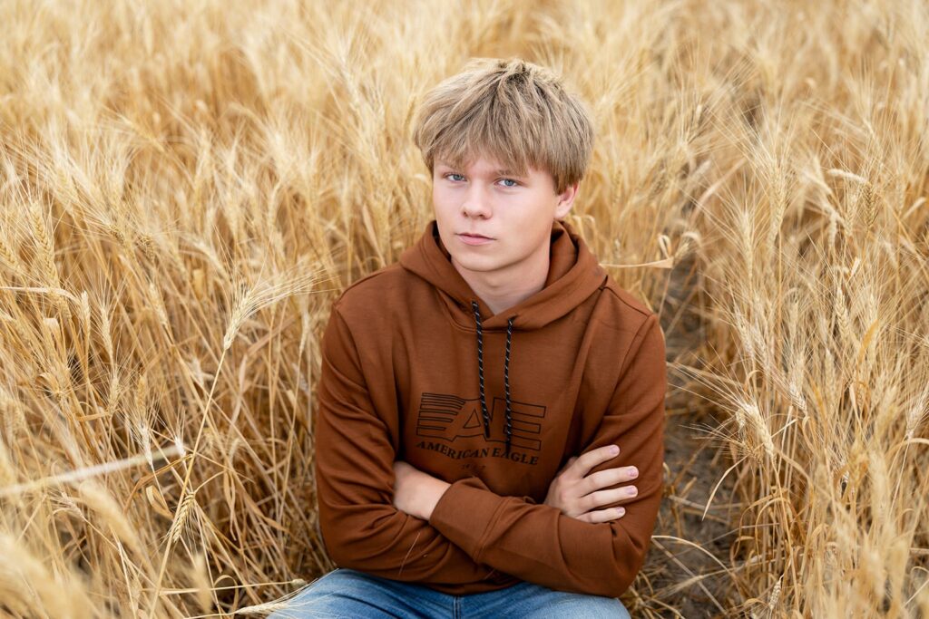 Teen boy sitting in golden wheat field during a travel session, wearing a rust-colored hoodie and jeans.
