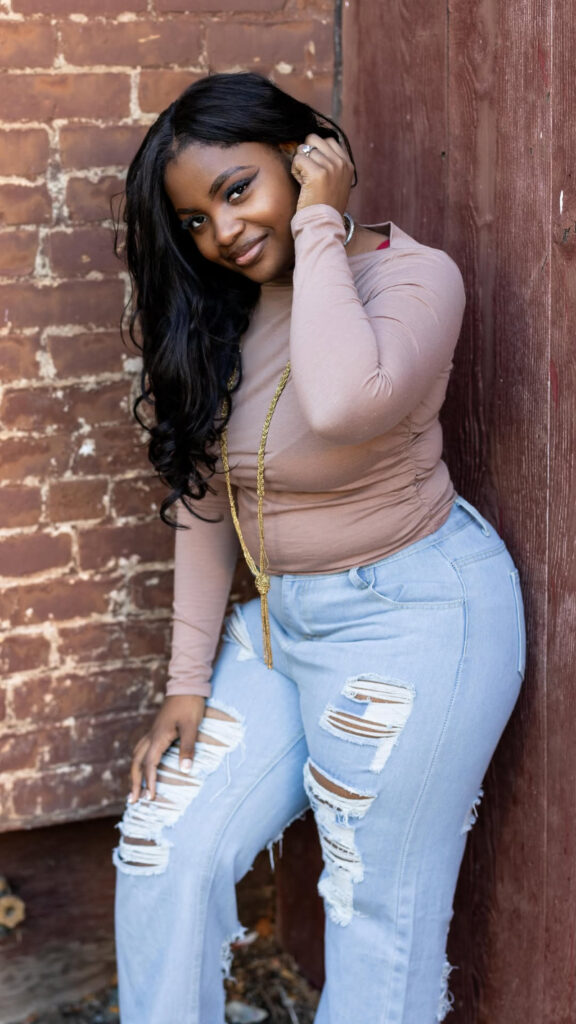 Confident woman posing against a rustic brick wall in casual ripped jeans and a long-sleeve top.