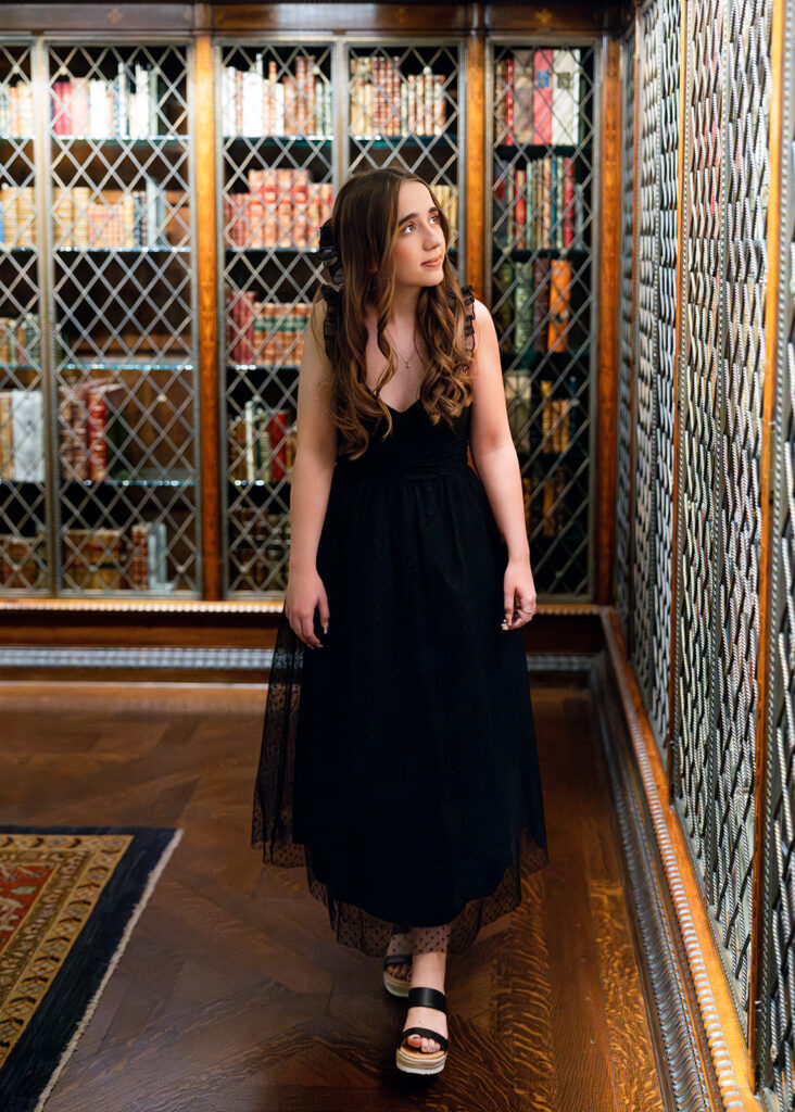 A young woman in a black dress walks amongst bookshelves during a NYC senior trip