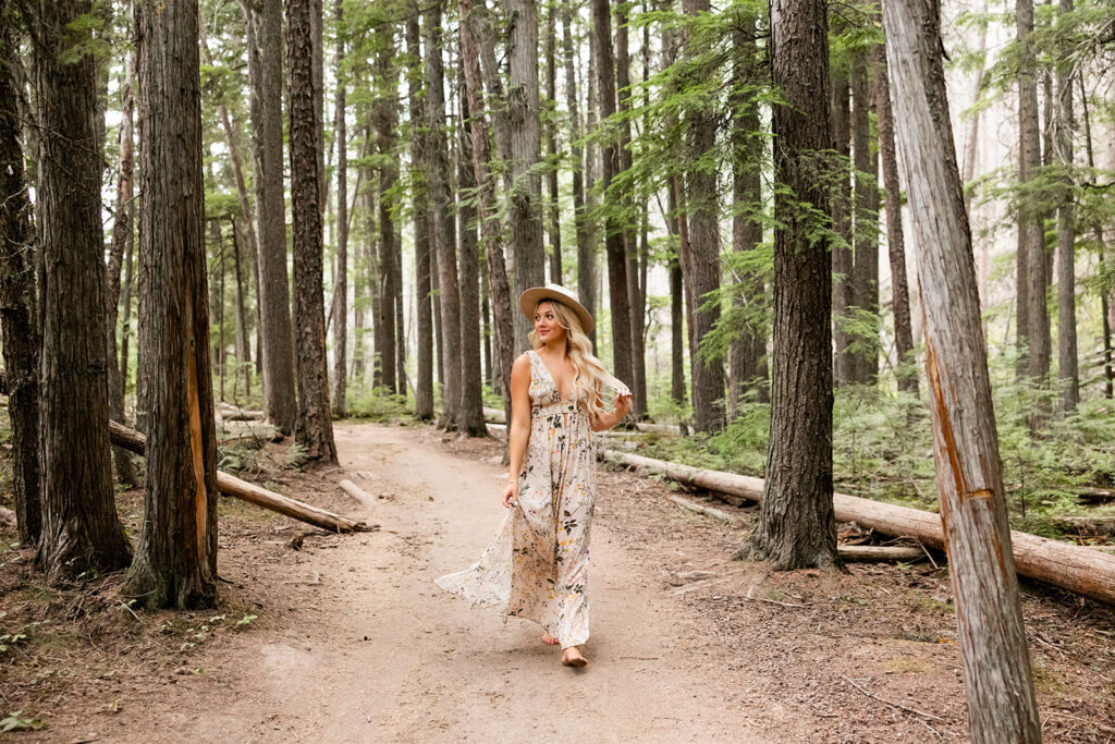 A young woman in a long floral dress walks through the forest at Glacier National Park during a senior photoshoot