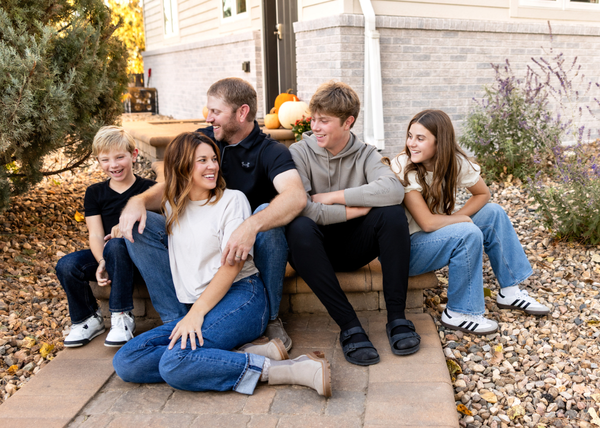 A joyful family of five sits on front steps at sunset, laughing together in cozy fall outfits outside their home. | imagen ai