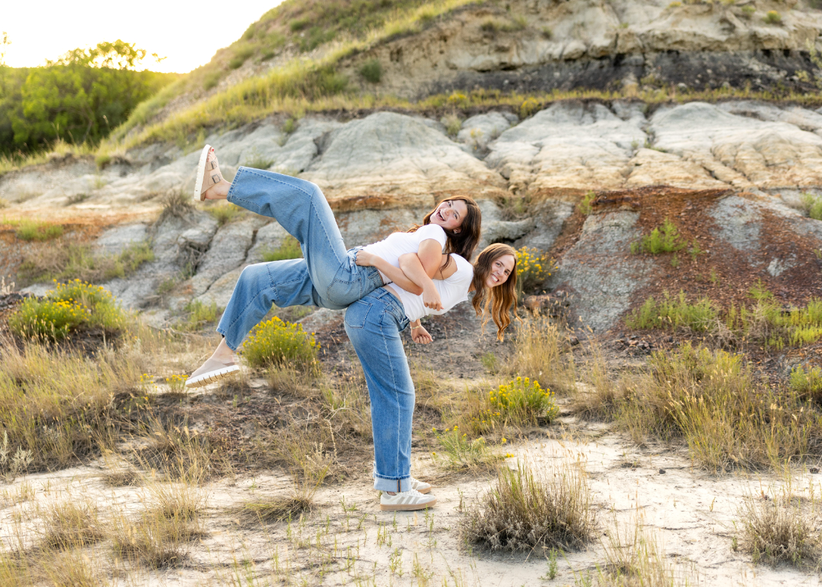 Two senior girls laugh in golden hills, one playfully lifting the other at sunset. Casual jeans, soft light, and genuine joy make it carefree and fun.