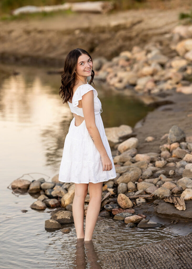 Senior girl in white dress smiling by water, natural light portrait on rocky shoreline