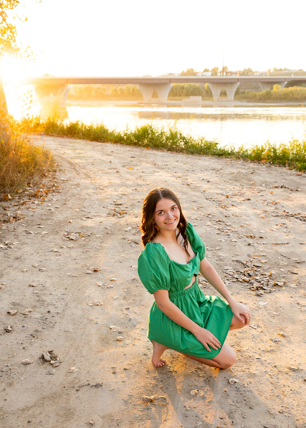 Senior girl in a bright green dress kneeling beside the river during sunset with soft backlighting and warm reflections on the water. This portrait captures the dreamy glow photographers love while showcasing golden hour photography tips for using natural light creatively.