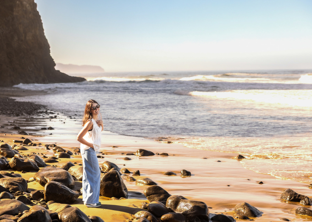 Senior portrait session on a rocky beach during sunset with soft ocean waves and warm golden light reflecting across the sand. The relaxed pose, natural movement, and dreamy coastal glow capture the feeling of carefree golden hour portraits.