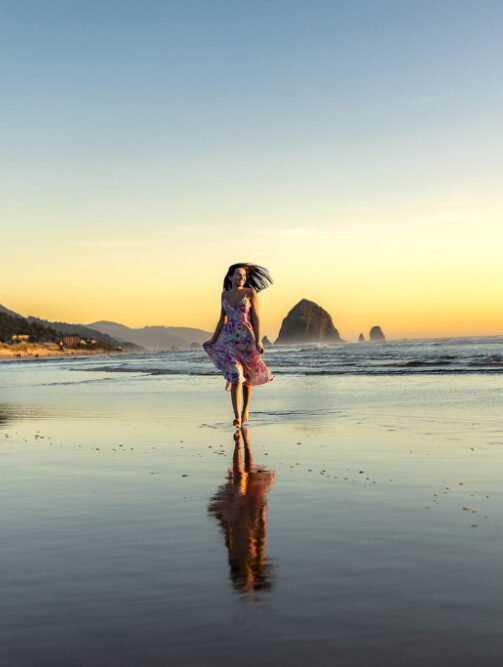 Young woman twirling barefoot along the shoreline during sunset with soft pastel skies and reflections in the wet sand. The golden light and open beach create a dreamy, movement-filled portrait with a natural and cinematic feel.