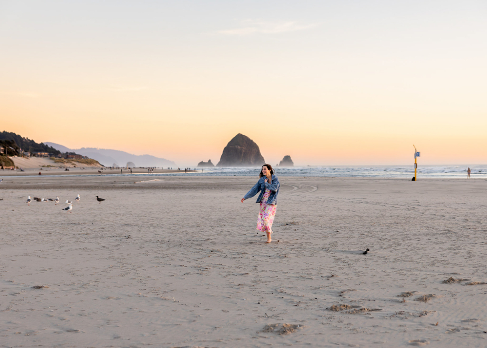 Senior girl walking barefoot on a wide beach at sunset with Haystack Rock in the distance and pastel skies overhead. The soft evening light, open space, and candid movement create a calm and airy coastal portrait atmosphere.