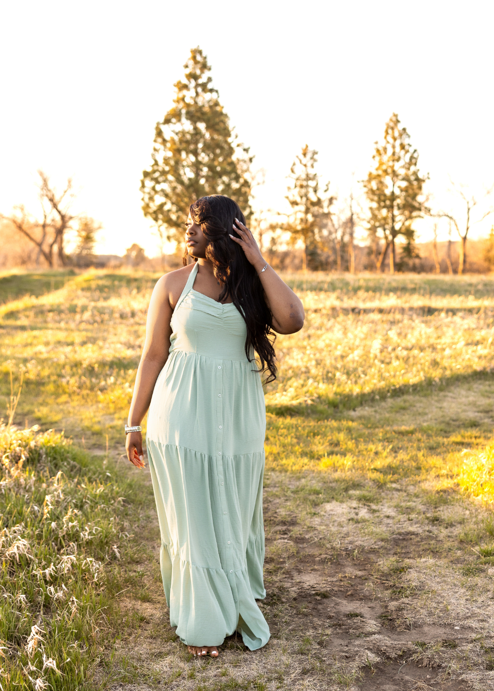 Outdoor portrait of a woman in a flowing sage green dress standing in a grassy field during golden hour. Backlighting from the setting sun creates soft highlights in her hair and warm tones across the landscape for a natural, glowing portrait.