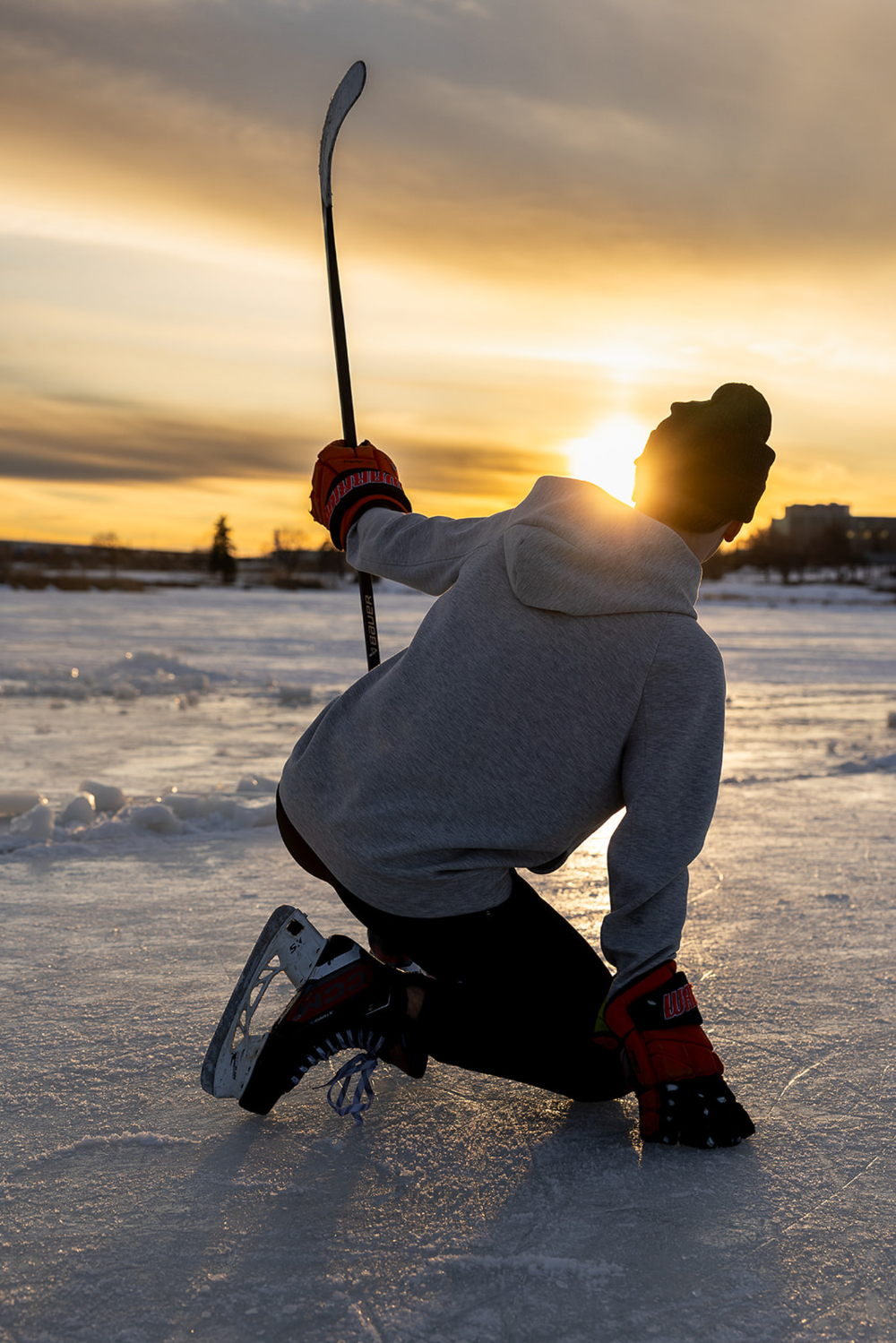 Hockey player kneeling on an outdoor ice rink at sunset with the sun glowing behind him. The dramatic silhouette, warm winter sky, and reflective ice create a bold athletic portrait that captures movement, strength, and golden hour light.