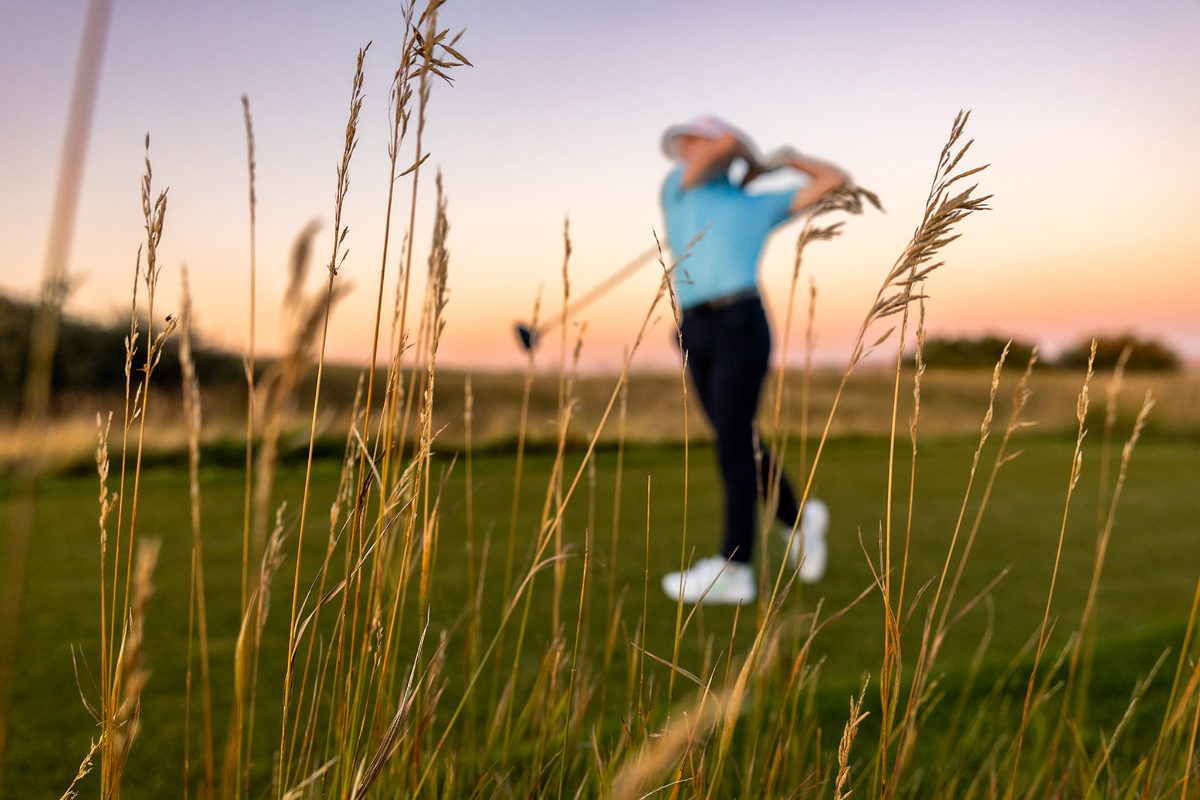 Creative foreground focus frames a golfer mid-swing at sunset, with tall prairie grasses sharp in the foreground and the athlete softly blurred behind. This image highlights storytelling through photography by capturing atmosphere, movement, and a fleeting moment rather than simply documenting the action.