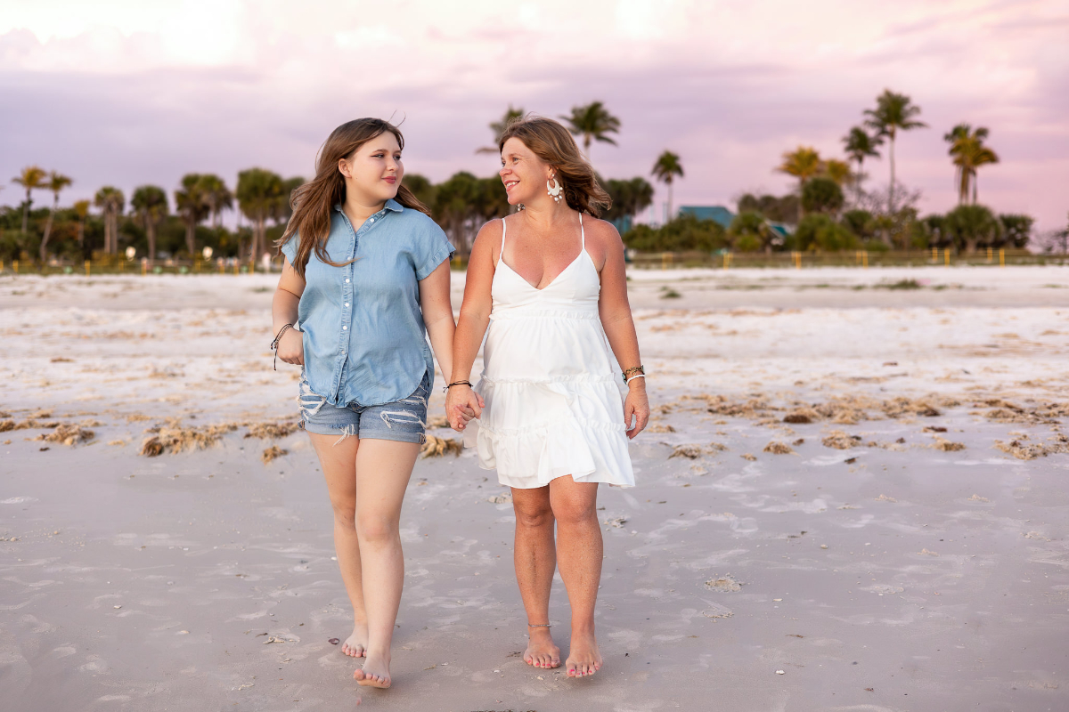 A mother and daughter walk hand in hand along a quiet beach beneath soft pink skies, sharing a genuine moment of connection. This image reflects storytelling through photography by preserving emotion, relationship, and the beauty of an everyday memory.