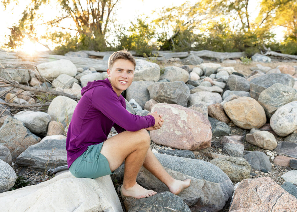 A senior sits among sunlit rocks near the water, relaxed and confident as golden light filters through the trees behind him. This image showcases storytelling through photography by incorporating environment, personality, and a sense of adventure into the portrait experience.