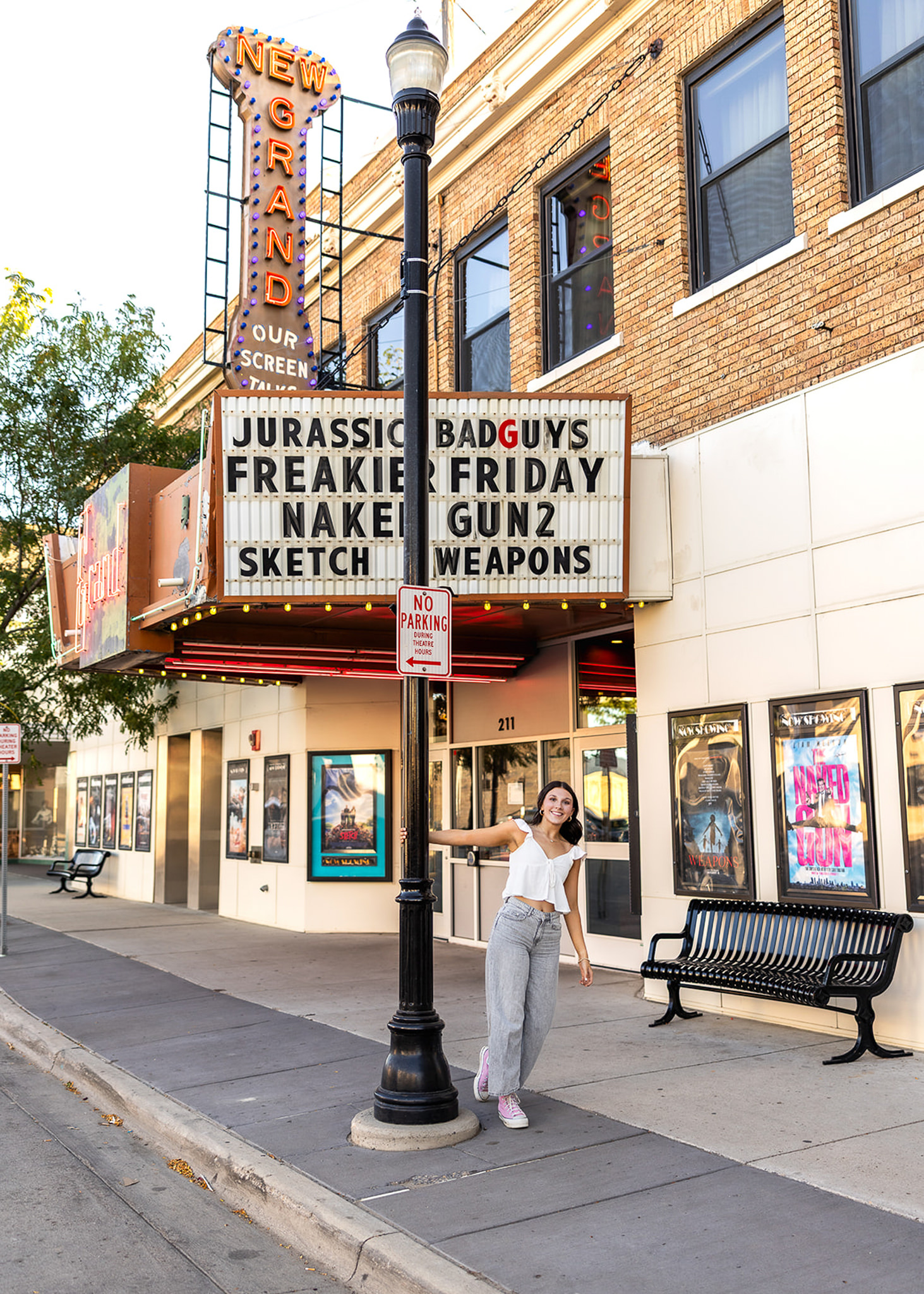 A high school senior smiles while posing beside a vintage streetlamp in front of a historic theater marquee downtown. The urban setting, bright signage, and playful pose create a fun, personality-filled senior portrait with a classic small-town feel.