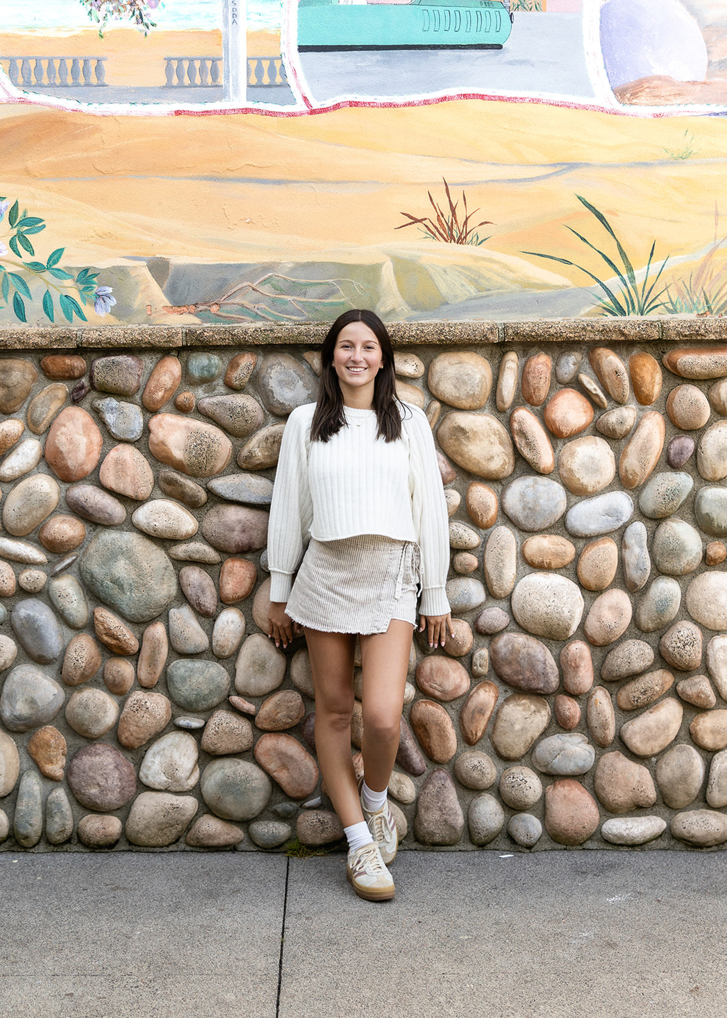 A smiling senior leans against a textured stone wall topped with a colorful mural. Neutral tones in her outfit complement the artistic backdrop, creating a modern senior portrait that highlights texture, color, and visual storytelling.