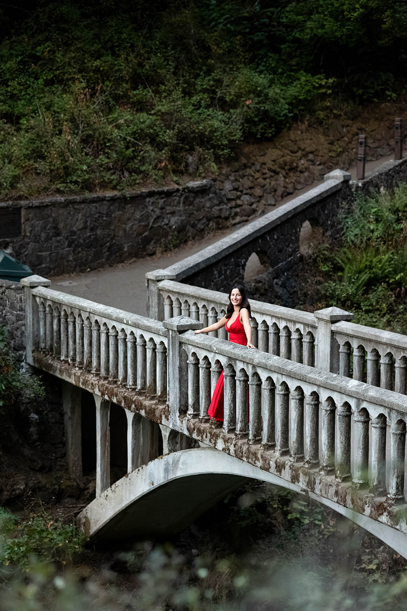 A senior in a flowing red dress stands on an elegant stone bridge surrounded by lush greenery. The elevated perspective and dramatic composition create a striking portrait that can inspire marketing ideas for photographers looking to showcase destination sessions.