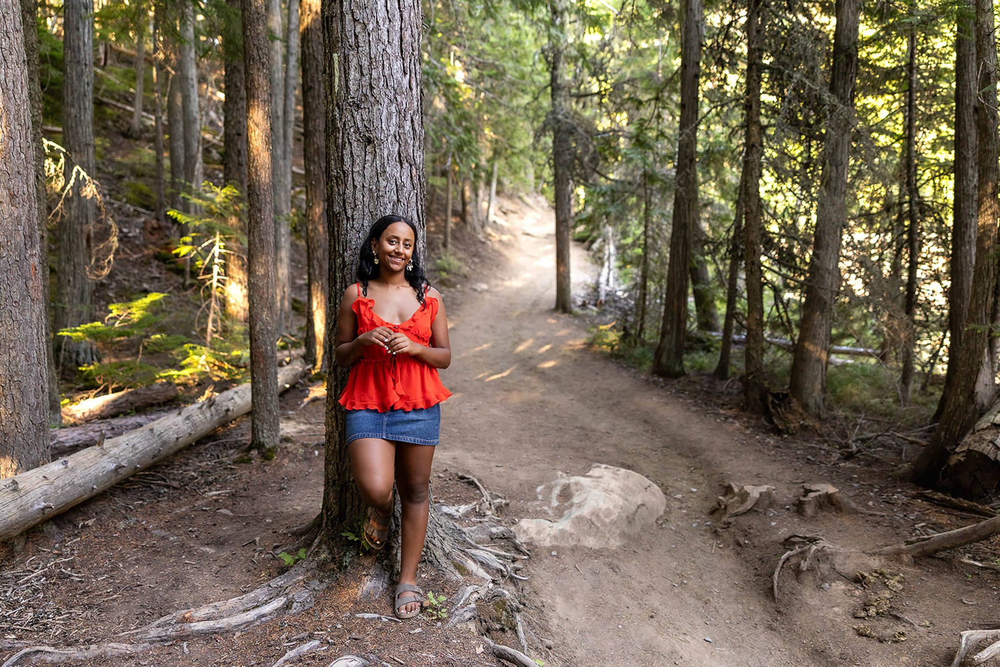 A senior stands against a tall tree along a forest trail surrounded by towering evergreens and soft afternoon light. This natural outdoor portrait showcases connection to nature and demonstrates creative marketing ideas for photographers who want diverse location options.