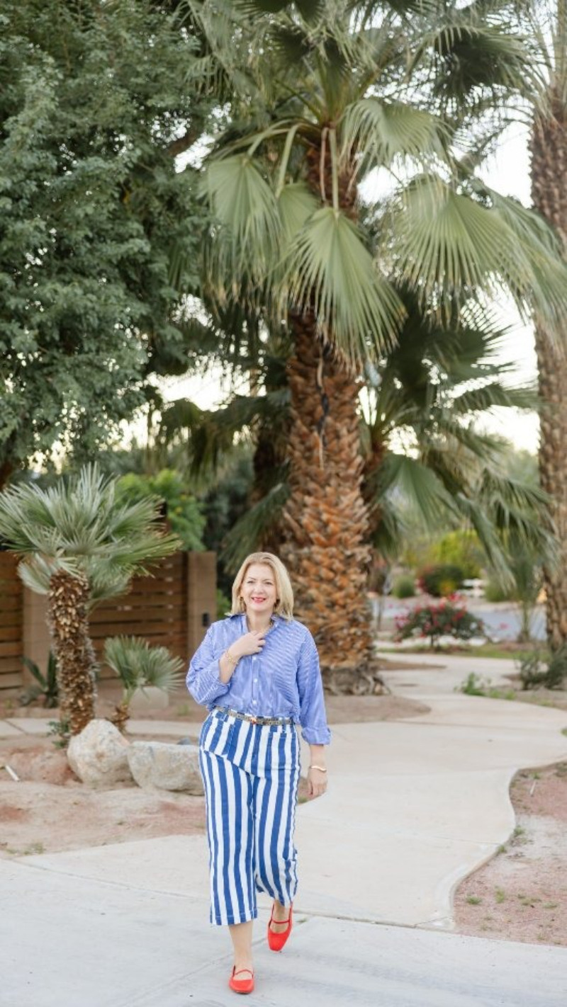 A woman walks along a palm-lined path in a desert setting, wearing blue striped pants and bright red shoes. The colorful wardrobe, relaxed movement, and destination backdrop create a memorable brand image that reflects how to stand out as a photographer.