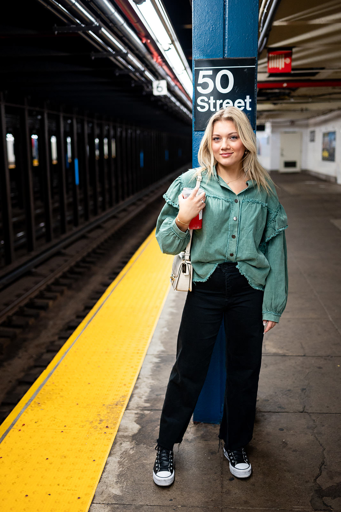 A blonde senior stands confidently on a New York City subway platform beneath a 50 Street sign, wearing a green blouse and black pants. The urban setting, leading lines, and natural expression create a bold destination senior portrait.