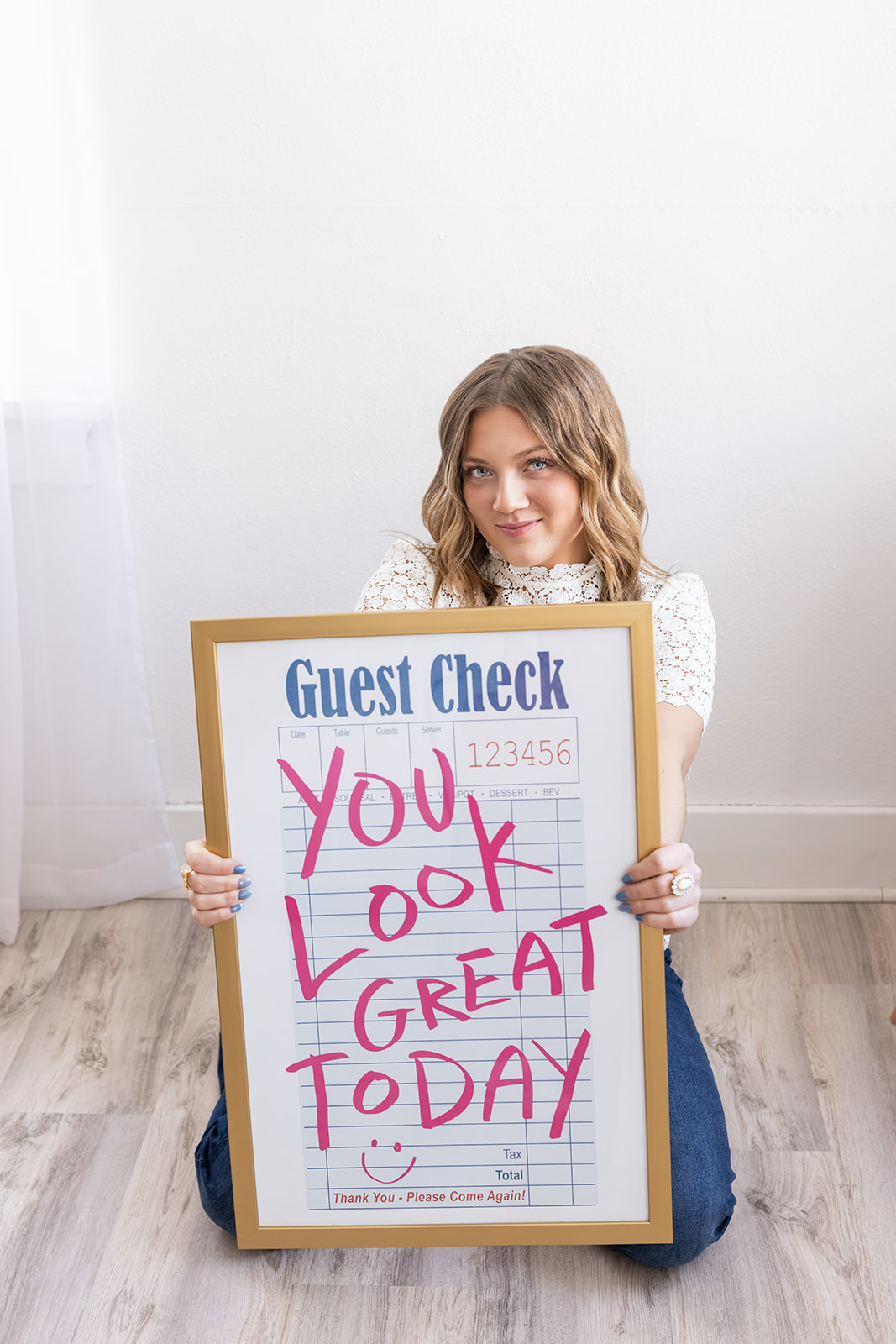 A young woman sits on a studio floor holding a framed sign that reads “You Look Great Today.” Bright natural light, clean styling, and encouraging messaging create a welcoming brand portrait perfect for entrepreneurs and creatives.