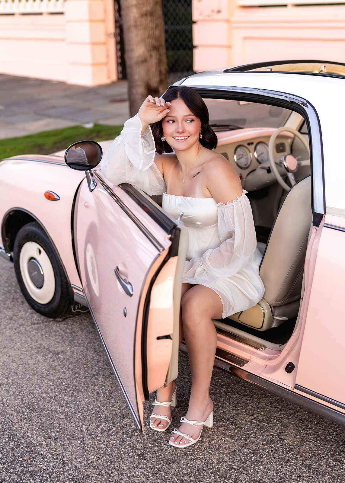 A senior girl in a flowing white dress sits in the doorway of a blush pink vintage convertible, smiling in soft golden light. This creative senior portrait showcases personality, styling, and storytelling—an example of how to stand out as a photographer.