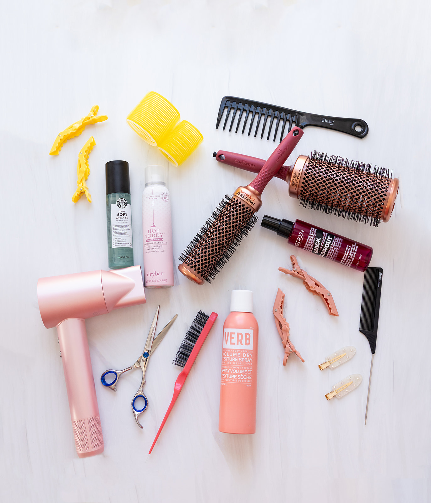 Flat lay of cosmetology tools arranged on a white surface during a branding photo shoot, featuring a pink hair dryer, styling brushes, shears, combs, rollers, clips, and professional hair products used to showcase a hairstylist's services and personality.