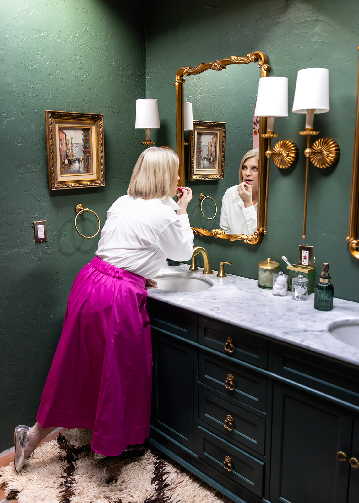 Woman applying lipstick in an elegant bathroom mirror during a branding photo shoot. Styled with a white blouse and bright pink skirt, the image captures an authentic getting-ready moment that adds personality and storytelling to a professional brand gallery.