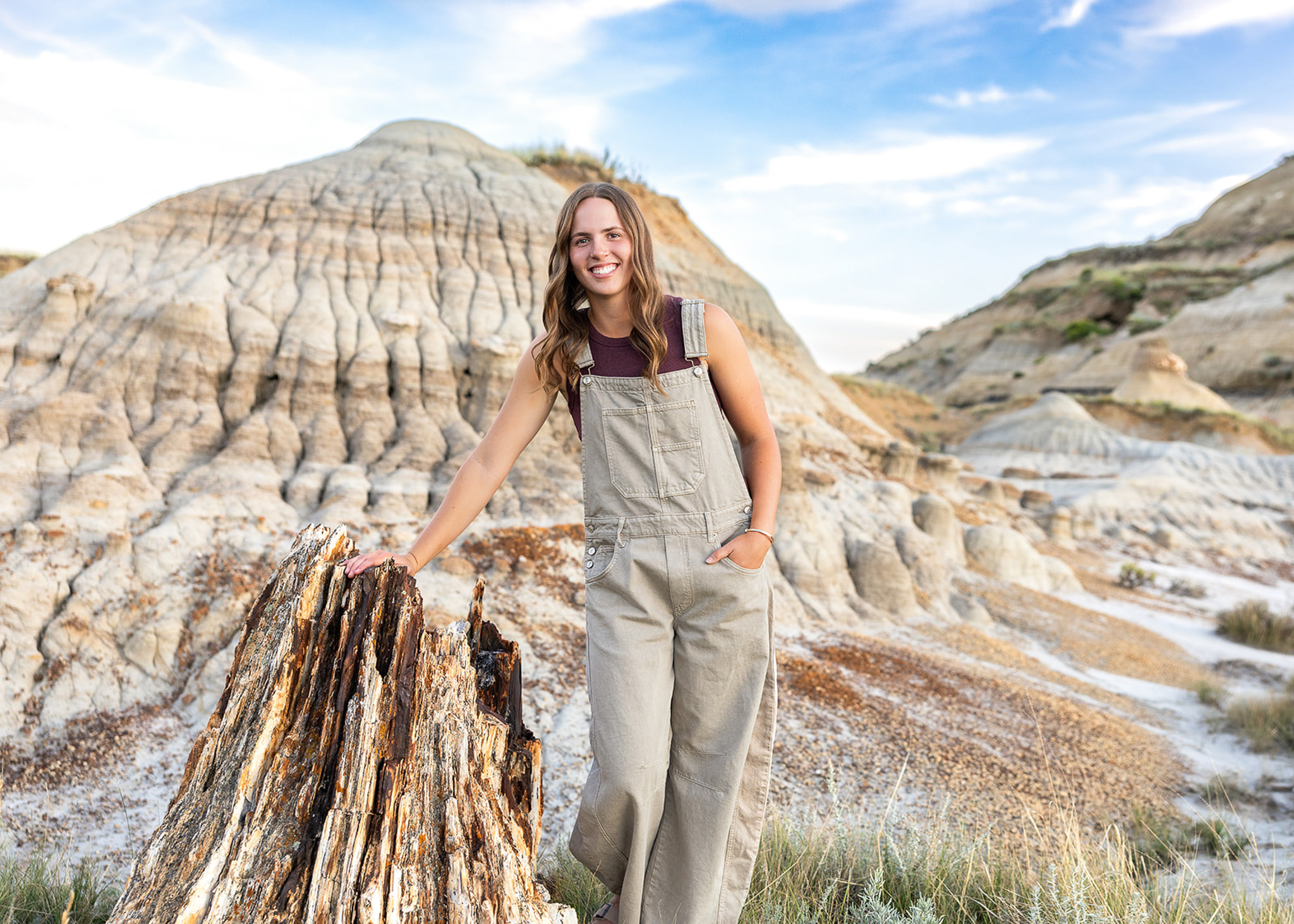 A high school senior stands beside a weathered tree stump in the North Dakota Badlands, smiling as layered rock formations stretch across the background. Soft evening light highlights the natural landscape and relaxed feel of the portrait.