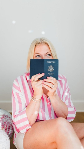 A blonde woman smiles while holding a U.S. passport in front of her face, hinting at upcoming travel adventures. Her pink striped shirt and bright, simple setting create a fun portrait celebrating a love for exploring new places.