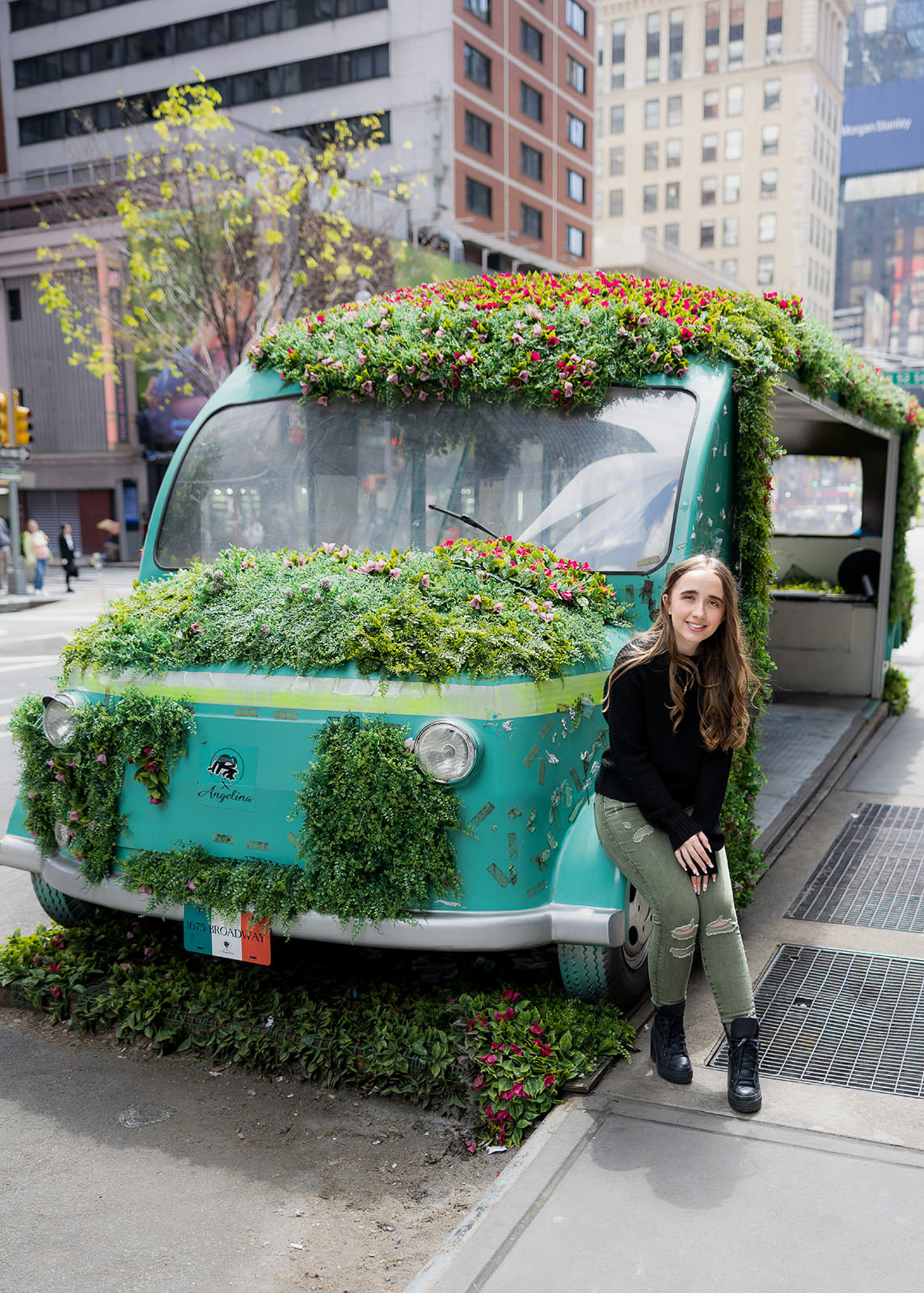 A high school senior smiles while sitting beside a vintage van covered in flowers on a busy New York City street. The colorful display and urban backdrop create a playful portrait that reflects the energy and charm of the city.